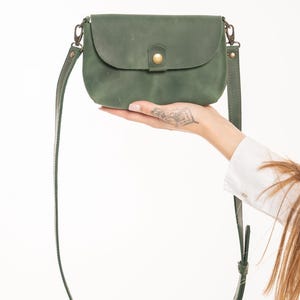 May include: A small, matte olive green leather crossbody bag with a rounded flap, a single brass-toned snap button closure, and a matching adjustable leather shoulder strap attached with brass-toned metal swivel clips on both sides.