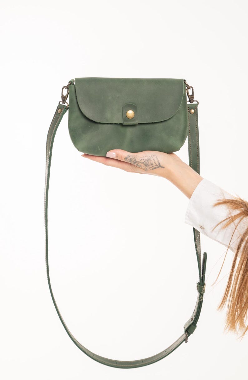 May include: A small, matte olive green leather crossbody bag with a rounded flap, a single brass-toned snap button closure, and a matching adjustable leather shoulder strap attached with brass-toned metal swivel clips on both sides.