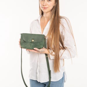 May include: A forest green crossbody bag with a rounded flap, a single brass-colored snap button closure, and a matching adjustable shoulder strap. The bag features a smooth, matte finish and is held horizontally in front of the body.