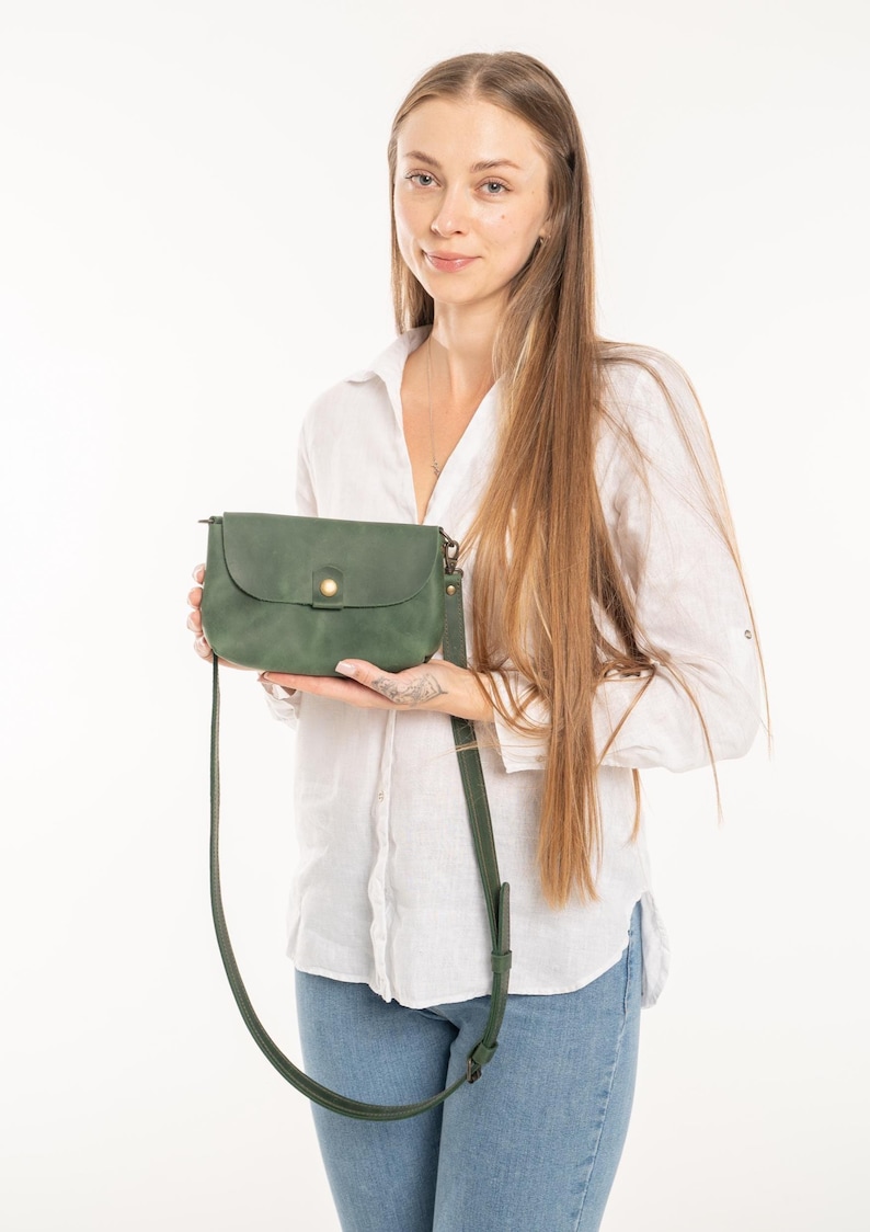 May include: A forest green crossbody bag with a rounded flap, a single brass-colored snap button closure, and a matching adjustable shoulder strap. The bag features a smooth, matte finish and is held horizontally in front of the body.