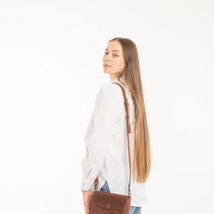May include: A brown leather crossbody bag with a rounded flap, a central metal snap closure, and a thin adjustable shoulder strap with metal hardware, worn over a white long-sleeved button-down shirt and blue denim jeans.