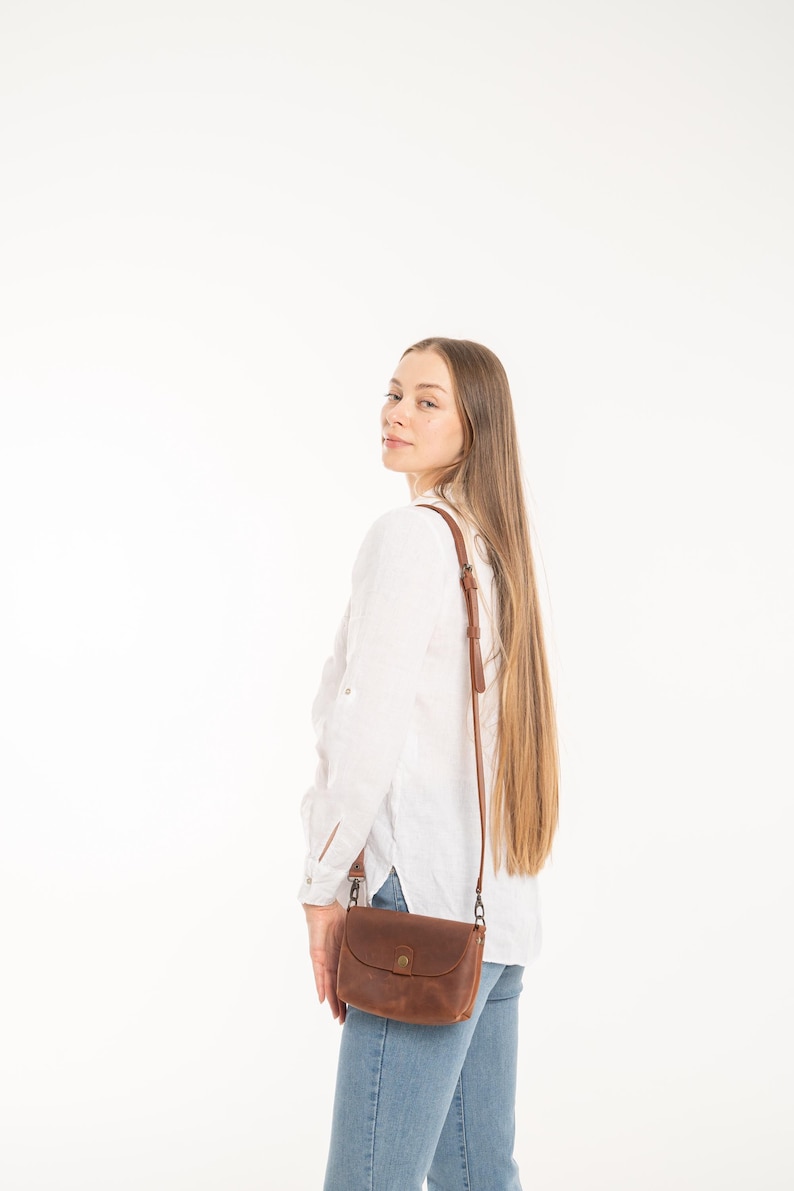 May include: A brown leather crossbody bag with a rounded flap, a central metal snap closure, and a thin adjustable shoulder strap with metal hardware, worn over a white long-sleeved button-down shirt and blue denim jeans.