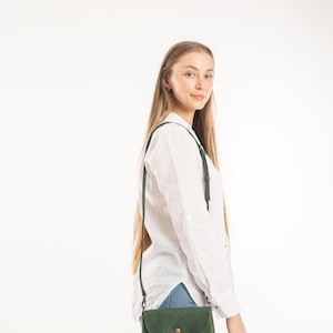 May include: A forest green leather crossbody bag with a rounded flap, a single brass-toned snap button closure, and a thin adjustable shoulder strap, worn over a white long-sleeved button-down shirt and light blue denim jeans.