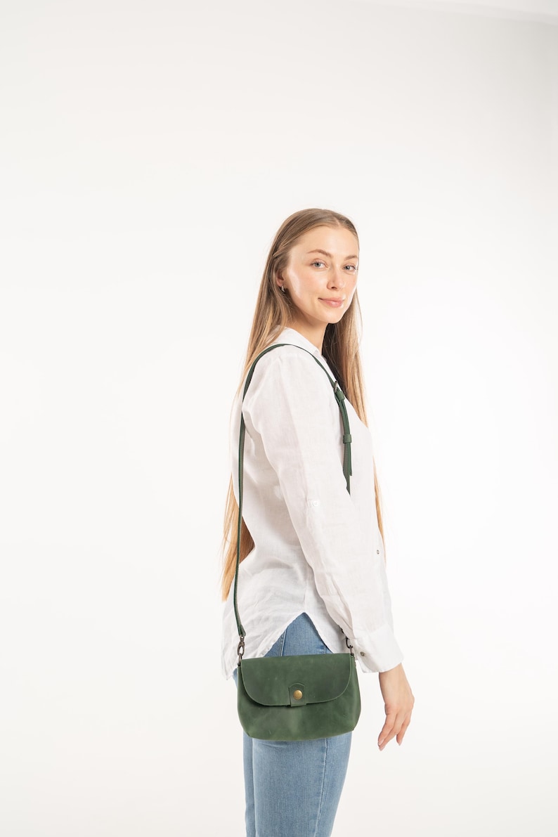 May include: A forest green leather crossbody bag with a rounded flap, a single brass-toned snap button closure, and a thin adjustable shoulder strap, worn over a white long-sleeved button-down shirt and light blue denim jeans.