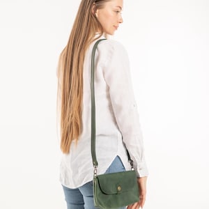 May include: A forest green leather crossbody bag with a long adjustable strap and a single snap button closure. The bag features a rectangular shape with rounded corners and is worn over the shoulder, resting at the hip with dark metal hardware.
