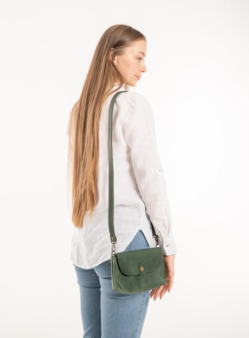 May include: A forest green leather crossbody bag with a long adjustable strap and a single snap button closure. The bag features a rectangular shape with rounded corners and is worn over the shoulder, resting at the hip with dark metal hardware.