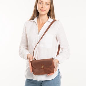 May include: A brown leather crossbody bag with a smooth matte finish, a rounded flap closure, and a single brass-colored metal snap button. The bag features a thin, adjustable leather shoulder strap attached to the sides with small metal hardware.
