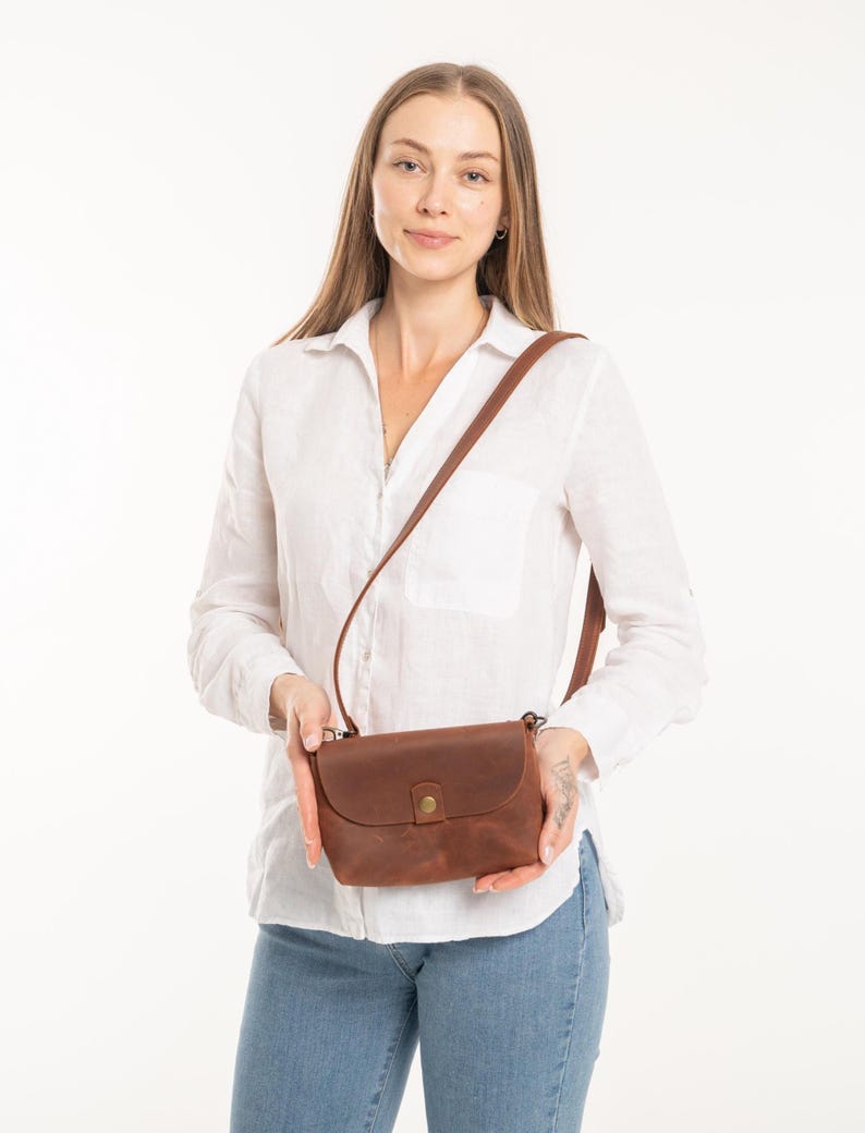 May include: A brown leather crossbody bag with a smooth matte finish, a rounded flap closure, and a single brass-colored metal snap button. The bag features a thin, adjustable leather shoulder strap attached to the sides with small metal hardware.