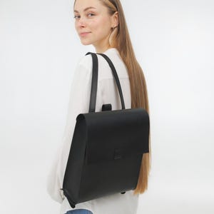 Puede incluir: Una mochila negra lisa con silueta rectangular, cierre de solapa plegable y dos correas verticales para los hombros. El bolso presenta un acabado liso y mate, con un diseño sencillo que incluye una lengüeta rectangular centrada en la solapa.
