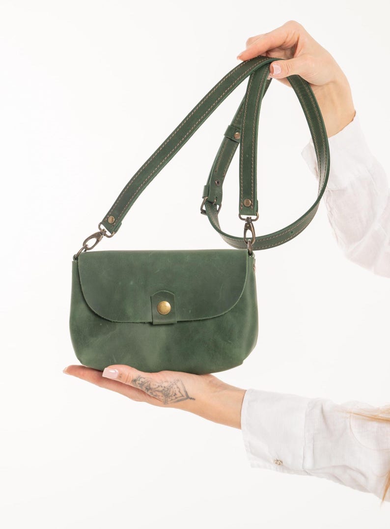 May include: A forest green leather crossbody bag with a rounded flap, a single brass-colored snap button closure, and an adjustable matching leather shoulder strap attached with metal swivel clips. The bag features visible stitching along the edges.