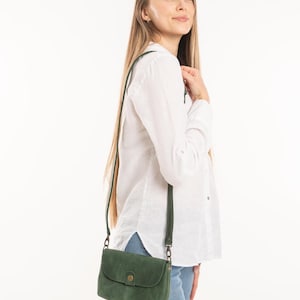 May include: A small, dark green leather crossbody bag with a rounded flap closure, a single brass-toned snap button, and a matching adjustable shoulder strap. The bag features a smooth, matte finish and is worn at the hip with a long, thin strap.