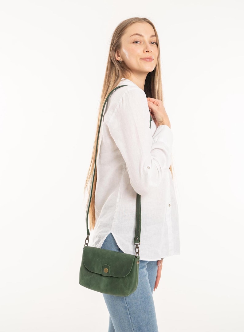 May include: A small, dark green leather crossbody bag with a rounded flap closure, a single brass-toned snap button, and a matching adjustable shoulder strap. The bag features a smooth, matte finish and is worn at the hip with a long, thin strap.