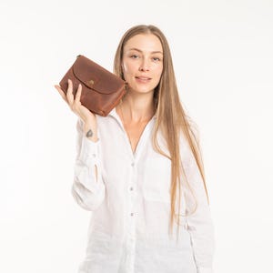 May include: A rectangular brown leather clutch bag featuring a curved flap closure secured with a single brass-colored metal snap button. The bag has a smooth, matte texture and visible stitching along the edges, presenting a simple and functional design.