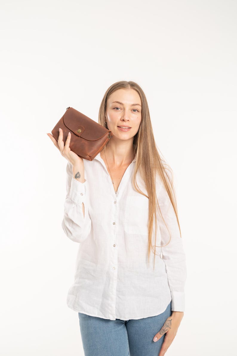 May include: A rectangular brown leather clutch bag featuring a curved flap closure secured with a single brass-colored metal snap button. The bag has a smooth, matte texture and visible stitching along the edges, presenting a simple and functional design.