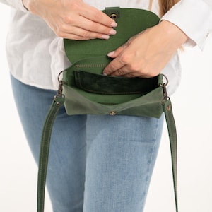 May include: A forest green leather crossbody bag with a flap closure, a single interior compartment, and a matching adjustable shoulder strap attached with dark metal hardware. The bag features visible stitching and a snap button fastener.