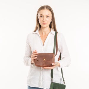 May include: A brown leather crossbody bag with a brass snap closure and a matching brown leather strap, held alongside a smaller dark green leather crossbody bag featuring a brass snap closure and a dark green leather strap.