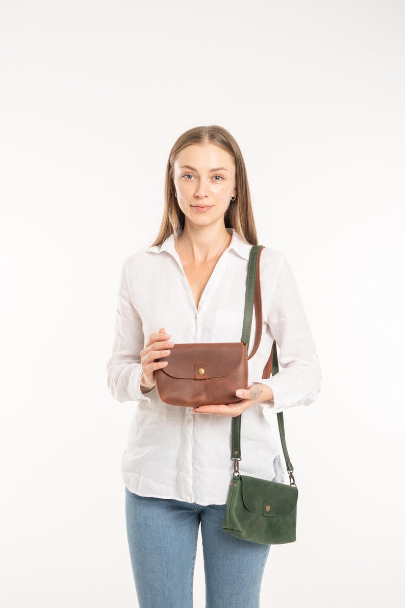 May include: A brown leather crossbody bag with a brass snap closure and a matching brown leather strap, held alongside a smaller dark green leather crossbody bag featuring a brass snap closure and a dark green leather strap.