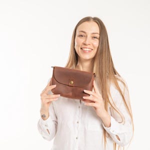 May include: A small, rectangular brown leather crossbody bag with a rounded flap closure. The bag features a single antique brass-colored metal snap button in the center of the flap and small metal rings on the sides for attaching a shoulder strap.