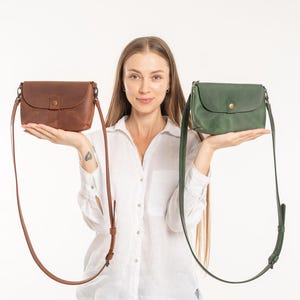 May include: Two leather crossbody bags, one in a rich brown and the other in a deep forest green. Each bag features a rounded flap, a single brass-toned snap button closure, and a long, adjustable matching leather shoulder strap with metal hardware.