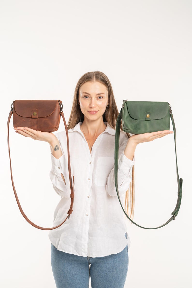 May include: Two leather crossbody bags, one in a rich brown and the other in a deep forest green. Each bag features a rounded flap, a single brass-toned snap button closure, and a long, adjustable matching leather shoulder strap with metal hardware.