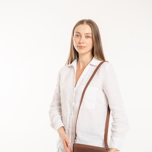 May include: A brown leather crossbody bag with a rounded flap, a single brass-colored snap button closure, and a thin adjustable shoulder strap. The bag features a smooth, matte finish and is constructed with visible stitching along the edges.
