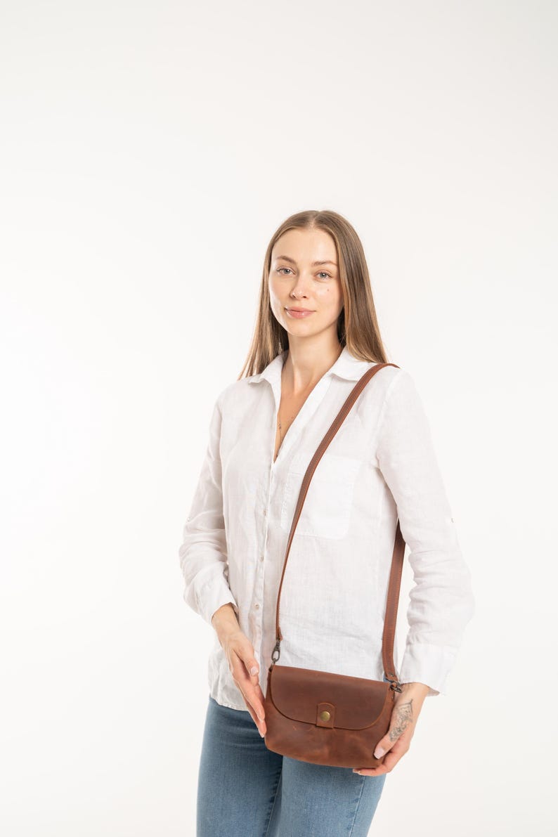 May include: A brown leather crossbody bag with a rounded flap, a single brass-colored snap button closure, and a thin adjustable shoulder strap. The bag features a smooth, matte finish and is constructed with visible stitching along the edges.