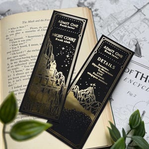 May include: Two black bookmarks with gold foil accents. The bookmarks feature a fantasy-themed design with a castle and mountains. The text on the bookmarks reads "ADMIT ONE Book lover" and "NIGHT COURT Book club". The other bookmark has the text "ADMIT ONE Book lover" and "DETAILS Location: Illyrian Mountains Time: 3AM Password: wingspan matters".