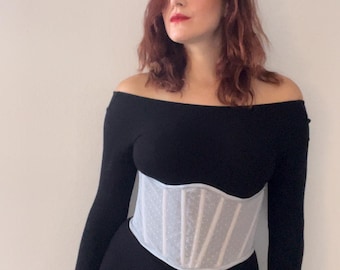 May include: A black off-the-shoulder long-sleeve dress with a white corset. The corset has a dotted pattern and a structured design. The dress is form-fitting, and the overall style is elegant and chic.