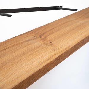 May include: A wooden shelf with a natural wood grain finish. The shelf is rectangular and appears to be made of solid wood. Above the shelf is a black metal bracket with two support arms, designed for wall mounting. The shelf is approximately 76 cm long.