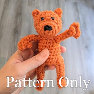 May include: Crochet pattern for a small orange bear with black eyes and nose. The bear is being held by a hand. The text "Pattern Only" is visible at the bottom of the image.