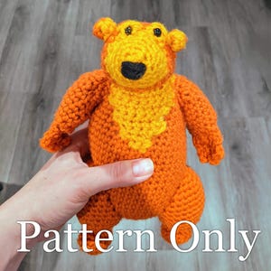 May include: Crocheted stuffed animal toy in the shape of a bear. The bear is orange with a yellow chest and black eyes. The bear is being held by a hand.