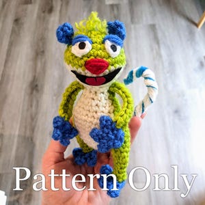 May include: A crocheted creature with a green and white body, blue paws and ears, and a red nose and mouth. The text "Pattern Only" is displayed at the bottom of the image.