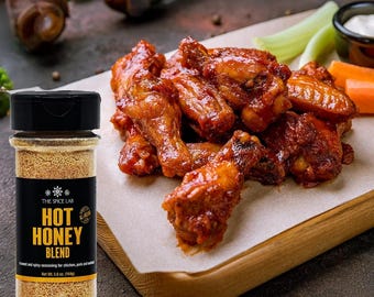 Könnte beinhalten: Ein Glas Hot Honey Blend-Gewürz neben einer Portion gekochter Chicken Wings. Die Wings sind mit einer rotbraunen Sauce glasiert und werden mit Sellerie, Karotten und einer kleinen Schüssel mit weißem Dip serviert. Das Glas hat ein schwarzes Etikett mit weißem Text.
