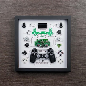 May include: A framed display of a disassembled PlayStation 4 DualShock 4 controller. The individual components are labelled and arranged on a white background. The controller is in the centre of the frame, with the words "DUALSHOCK 4" printed below it.