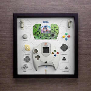 May include: A framed artwork featuring a disassembled Sega Dreamcast controller. The controller is white with a black rubber pad and a green, red, and blue button. The frame includes a circuit board with the text "SEGA" and "Sonic Adventure" printed on it.