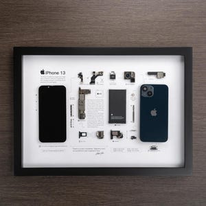 Op de afbeelding: Een witte ingelijste print met een zwarte iPhone 13 op een witte achtergrond. De print toont de componenten van de telefoon, inclusief de camera, batterij en het display. De tekst "There's a phone in Buddhism. Beginners mind. It's wonderful to look at a beginner's mind." is onder de telefoon afgedrukt.