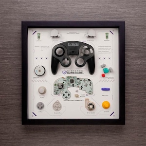 Könnte beinhalten: Eine gerahmte Darstellung der zerlegten Teile eines Nintendo GameCube-Controllers. Die Darstellung umfasst die Controller-Hülle, die Tasten und die Platine. Der Text "Nintendo GameCube" ist auf der Darstellung aufgedruckt.