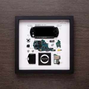 Może przedstawiać: Oprawiony wyświetlacz rozmontowanej konsoli do gier Sony PSP 1000. Konsola jest rozłożona na białym tle, a każdy element jest opisany. Wyświetlacz zawiera płytę główną konsoli, przyciski, ekran i inne części.