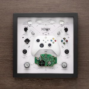 Könnte beinhalten: Ein weißer Xbox-Controller, zerlegt und auf einem weißen Hintergrund mit dem Text "XBOX" und "LOGIC BOARD ASSEMBLY" ausgelegt. Der Controller ist von seinen Einzelteilen umgeben, darunter Knöpfe, Trigger und die Platine.