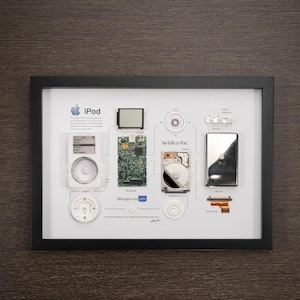 May include: A framed display of the disassembled parts of an original iPod, including the hard drive, circuit board, screen, and buttons. The text "Say hello to iPod" and "1,000 songs in your pocket" are visible.
