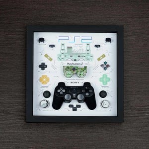 May include: A framed display of a PlayStation 2 motherboard and controller. The motherboard is green and has the text "PlayStation 2" printed on it. The controller is black and has the text "SONY" printed on it.