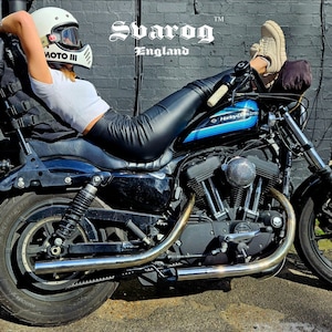 May include: A black Harley Davidson motorcycle with a blue stripe on the gas tank. A person is sitting on the motorcycle with their legs extended and wearing a white helmet with a black visor and black leather pants. The motorcycle is parked in front of a brick wall with the text "Svarog England" on it.