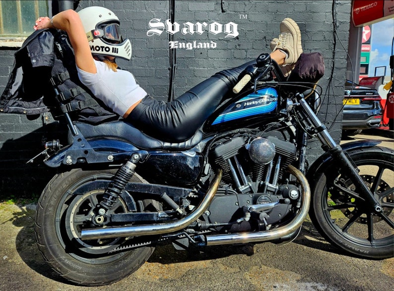 May include: A black Harley Davidson motorcycle with a blue stripe on the gas tank. A person is sitting on the motorcycle with their legs extended and wearing a white helmet with a black visor and black leather pants. The motorcycle is parked in front of a brick wall with the text "Svarog England" on it.
