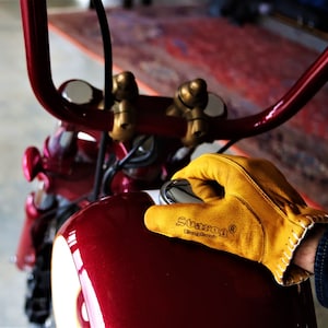 May include: A close-up of a red motorcycle gas tank with a yellow leather glove resting on top. The glove has the text "SUGAR&reg;  Motorcycle" printed on it.