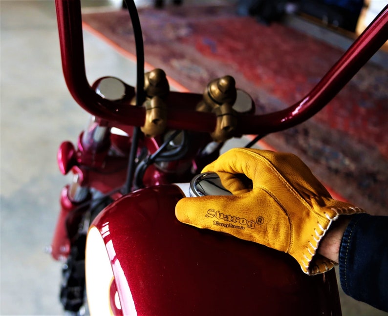 May include: A close-up of a red motorcycle gas tank with a yellow leather glove resting on top. The glove has the text "SUGAR&reg;  Motorcycle" printed on it.