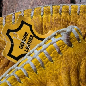May include: Close-up of a pair of yellow leather gloves with white stitching. A tag that reads "GENUINE LEATHER" is attached to the gloves.