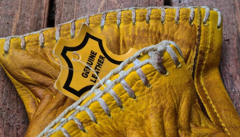 May include: Close-up of a pair of yellow leather gloves with white stitching. A tag that reads "GENUINE LEATHER" is attached to the gloves.