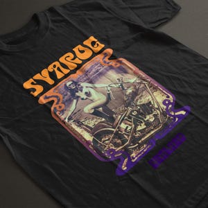以下が含まれることがあります： 黒いTシャツ。レトロなバイクに乗る人のグラフィックが特徴です。「SYARGE」というオレンジ色の文字と、紫とオレンジのデザインが含まれています。柔らかく、暗い色の生地で作られています。