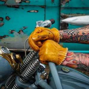 May include: A pair of brown leather motorcycle gloves with white stitching. The gloves have the word "Durston" embossed on the back of the left glove. The gloves are worn by a person with heavily tattooed arms.