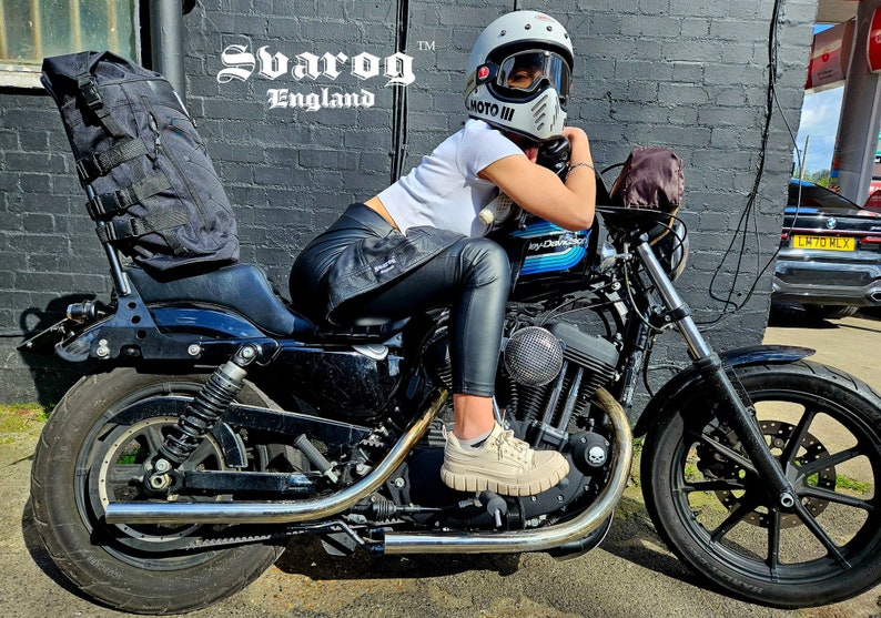 May include: A woman wearing a white shirt and black leather pants sits on a black motorcycle. She is wearing a white helmet with a black visor and a black and white motorcycle jacket. The motorcycle has a black leather seat and a black and chrome exhaust pipe. The motorcycle is parked in front of a brick wall with the words "Svarog England" written on it.